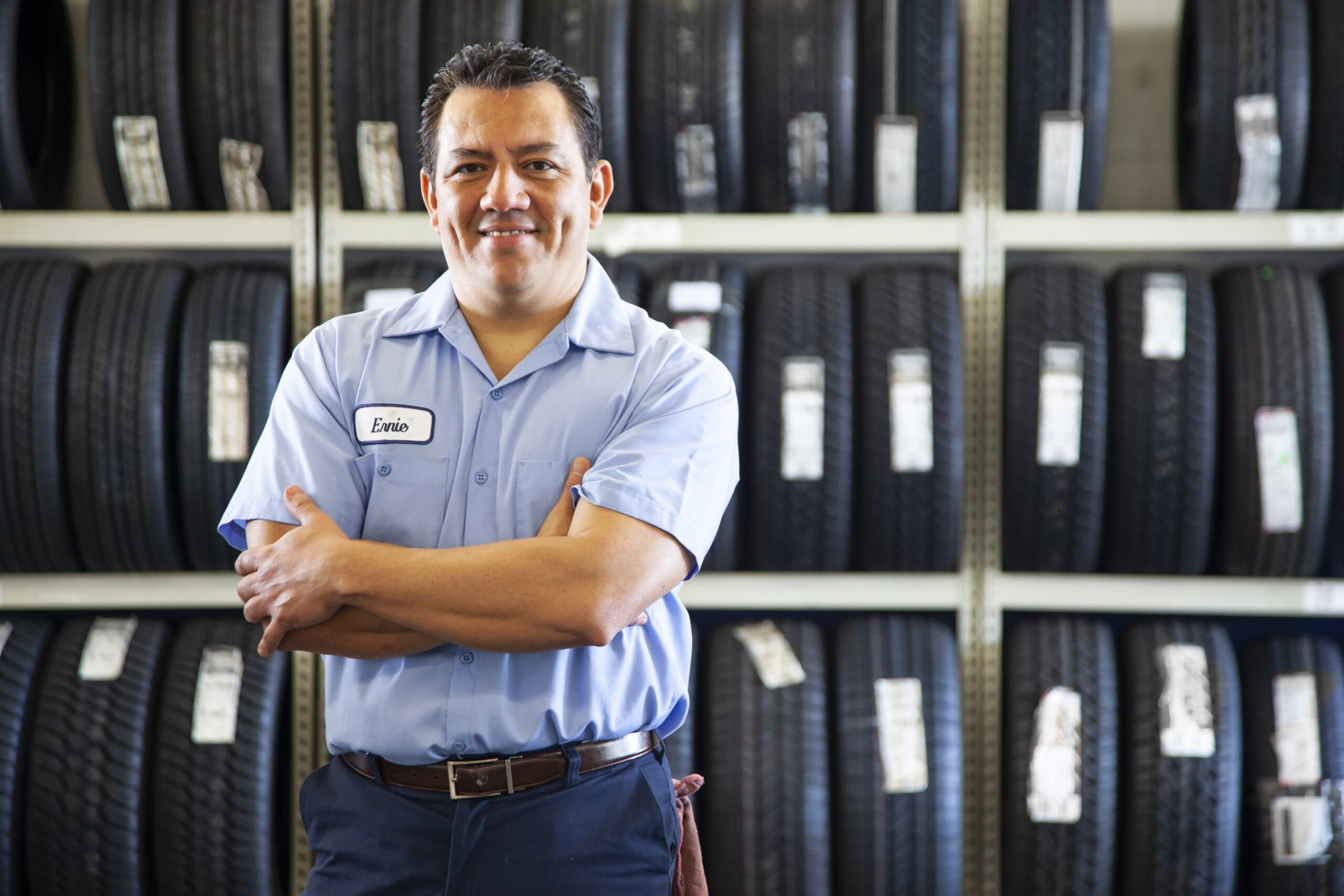 Homem com uniforme azul em frente a pneus organizados em prateleiras, representando dono de loja de pneus.