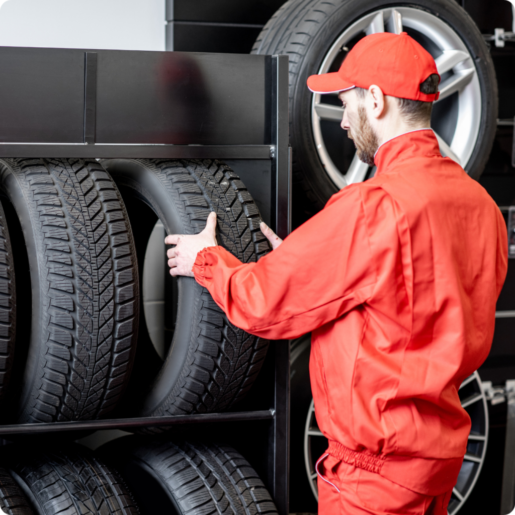 Profissional da Distribuidora Pneu Forte em uniforme vermelho organizando pneus novos em estante metálica, representando agilidade no atendimento e estoque disponível.